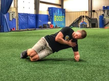 Dr. Aaron Horschig apoiado de lado pelos joelhos e cotovelos em chão de grama sintética em posição do side plank